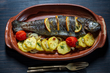 Baked trout fish with potato slices, lemon, tomato and spices on a clay plate, closeup