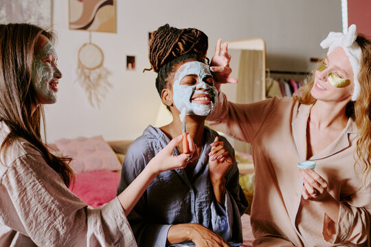 Medium shot of African American young woman with bun of dreadlocks wrinkling face while her biracial friends applying clay mask on her face - Powered by Adobe