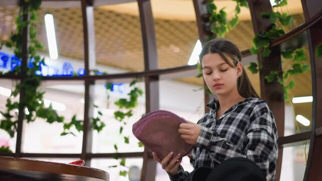 Exhausted girl sits at wooden table, placing mirror down, appearing stressed yet determined, reen plants and warm indoor lighting create a cozy and introspective atmosphere
