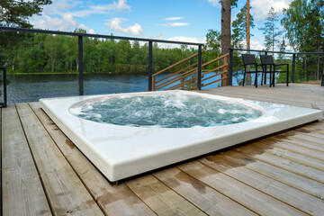 Modern hot tub embedded in a wooden terrace with a scenic view of a peaceful summer lake surrounded by lush green forest. Perfect spot for relaxation and enjoying nature in comfort.