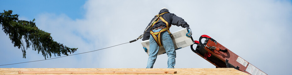 Fototapeta premium Workman in safety harness on wood framed house roof carrying package of roofing materials delivered by conveyor belt on a sunny winter day, home development construction project 