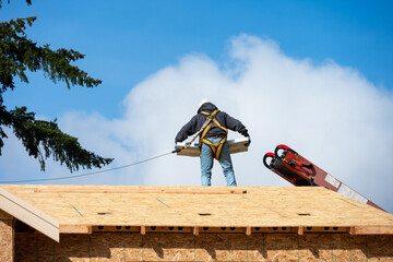 Workman in safety harness on wood framed house roof carrying package of roofing materials delivered...