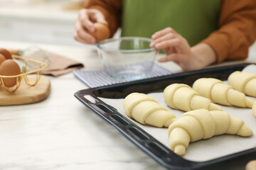 Woman breaking egg into bowl near raw croissants at white marble table indoors, selective focus