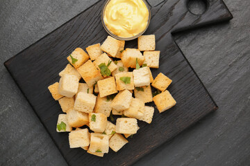 Tasty crispy croutons with cut parsley and sauce on dark textured table, top view