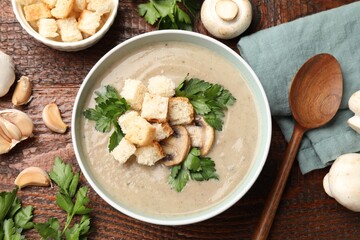 Delicious mushroom cream soup with croutons, parsley, ingredients and spoon on wooden table, flat lay