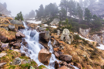 Waterfall cascading down rocky slope, misty mountain setting. Serene mountain waterfall flowing through rocks in a misty forest with snowy patches. GR11, Restanca, Lleida