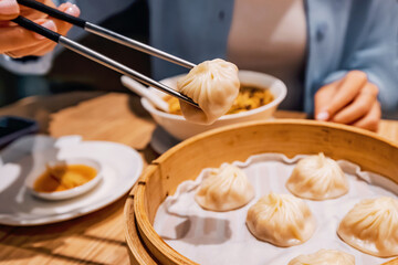 Metal chopsticks holding a xiaolongbao dumpling over a bamboo steamer basket in a restaurant