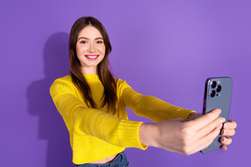 Naklejka premium Smiling young woman in casual yellow sweater taking a selfie against a violet background showcasing cheerful and trendy lifestyle