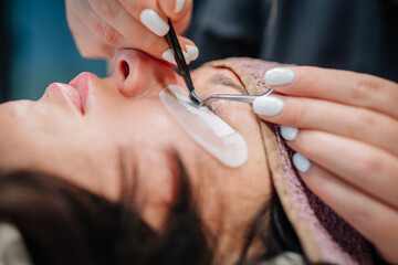 Extreme close-up of a beautician applying individual eyelash extensions with tweezers to a client’s eye, using under-eye pads for protection..