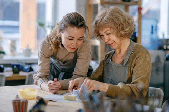 Teacher guides an elderly student, demonstrating sculpting techniques in a hands-on class. Senior woman practices clay modeling with help from a younger instructor in a ceramics studio.