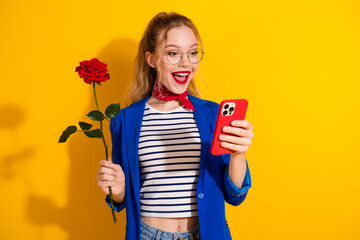 Young woman wearing glasses holding a rose while checking her smartphone against a vibrant yellow backdrop