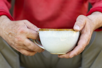 A close-up of rough, weathered hands holding a rustic ceramic cup filled with hot tea. The person is wearing a red jacket, and the warm, earthy tones create a cozy and authentic atmosphere. 