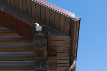 A security camera is mounted on the corner of a wooden house under a brown metal roof. The camera is directed outward, ensuring surveillance and protection. 