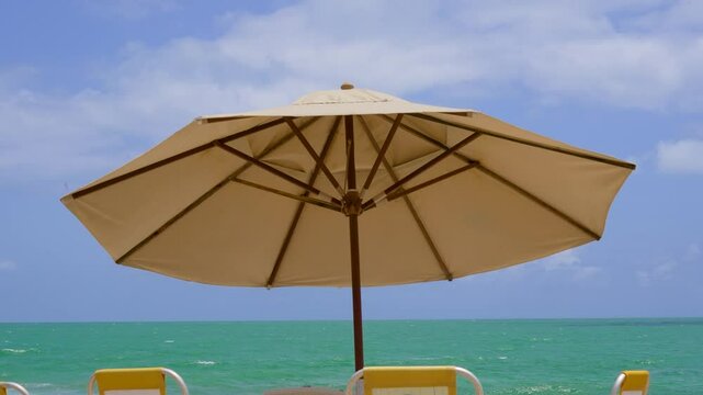 Relaxing beach scene with a large beige umbrella shading yellow lounge chairs, facing the turquoise ocean under a bright blue sky. A peaceful tropical getaway for summer vacations.