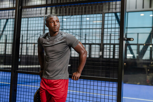 Athletic man in sportswear leaning on padel court fence, looking determined