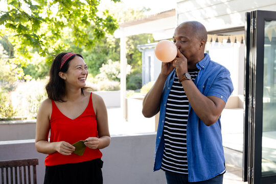 At party, African American man blowing balloon while woman in red top smiling on deck