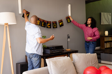 Two diverse men decorating living room with welcome banner for celebration, at home