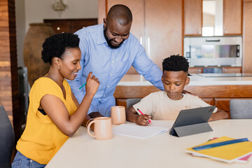 Family helping child with homework at kitchen table, using tablet and notebooks