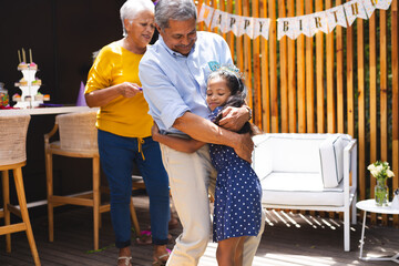 At party, celebrating birthday in garden, grandfather hugging granddaughter with family around them