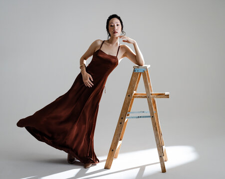 Elegant asian woman in flowing brown dress posing gracefully with wooden ladder
