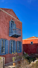 Traditional Greek House With Blue Shutters And Balcony, Rustic Red Walls, Sunny Day With Clear Blue Sky, Taken In Greece During Summer, Ideal For Travel, Architecture, And Cultural Heritage Concepts