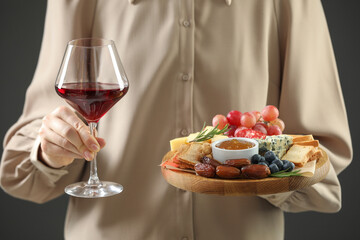Woman holding board with different types of delicious cheese, other snacks and wine on grey background, closeup