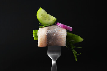 Fork with marinated herring fillet, cucumber, rosemary and onion ring on black background, closeup