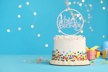 Delicious bento cake with colorful sprinkles and topper with words Happy Birthday on light blue table against blurred lights. Space for text