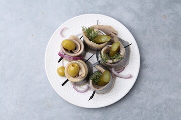 Delicious herring rolls with olives, pickles, dill and onion on gray textured table, top view