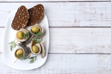 Delicious herring rolls with olives, dill and bread on white wooden table, top view. Space for text
