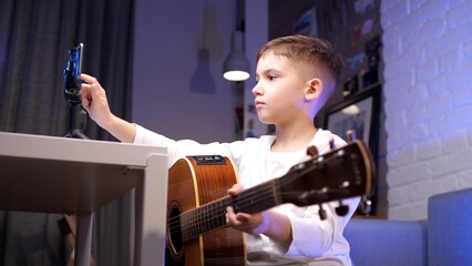 Young boy is sitting on a couch holding an acoustic guitar while adjusting his smartphone which is recording a video blog