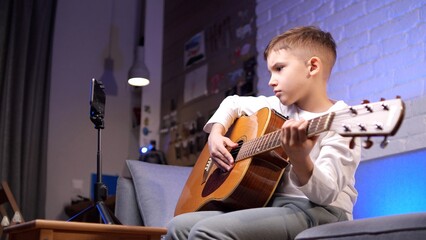 Obraz premium Young boy sits on a cozy couch, playing an acoustic guitar and recording himself with a smartphone, showcasing his passion for music