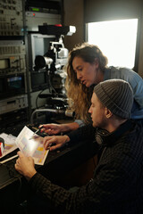 Young woman pointing at paper document during discussion of broadcast schedule with male operator while preparing for shooting