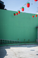 Red pineapple-shaped lanterns hanging against a green wall, with string lights intertwined between them, and a ladder lying on the ground