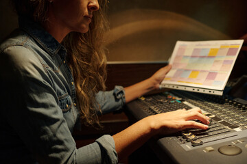Cropped shot of young female journalist with broadcast schedule in hand adjusting sound while moving knobs on console
