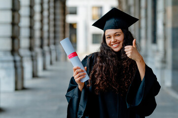 Young latin woman wearing graduation gown holding diploma and showing thumb up, celebrating...
