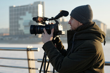 Side view of smiling young man in warm jacket and beanie adjusting video camera while preparing for...
