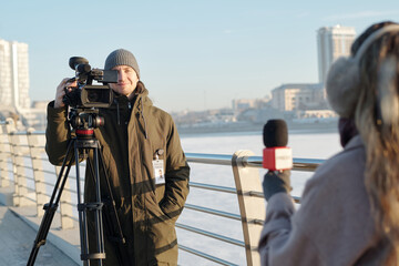 Happy young man shooting news reportage on professional video camera while standing in front of...