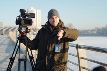 Young man with professional video camera consulting reporter while preparing for filming news or...