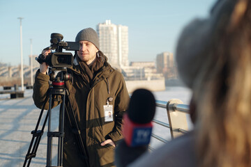 Focus on young male operator or videographer shooting female reporter with microphone while...