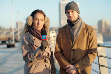 Young man standing next to female reporter of TV channel with microphone while both looking at camera