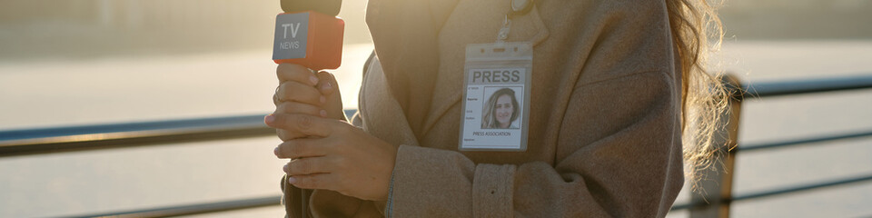 Header of hands of female reporter with microphone standing in front of camera against riverside in...