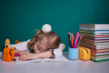 Young girl in school uniform falls asleep on her notebook at a desk filled with books and pencils, indicating exhaustion or fatigue during study time in a classroom setting.
