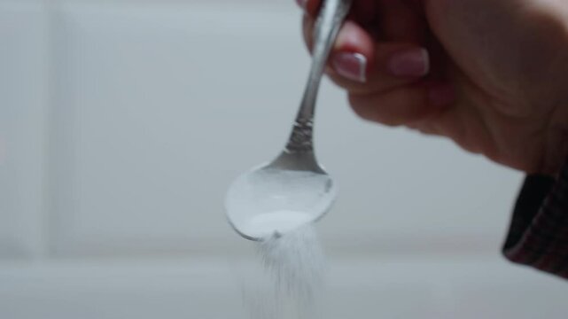 Close-up of hand holding spoon as sugar is gradually poured with bright tiled background, the fine sugar particles fall delicately, creating a soft culinary moment in a well-lit kitchen setting