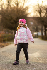 Fototapeta premium A little girl in a pink jacket and hat joyfully runs on a paved path in a park during golden hour. Chalk hopscotch is visible on the ground, with blurred trees and warm sunlight in the background.