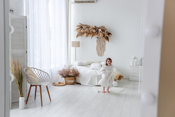 Cheerful little girl with long brown hair wearing a white dress is running barefoot in a cozy white bedroom with bohemian decoration, playing hide and seek