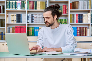 Young male teacher working in library sitting at desk with laptop books