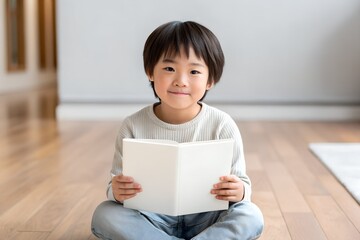 Asian child holding blank white book mockup while sitting on floor. Clean book cover template for children’s literature or educational content.