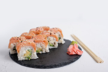 Plate of fresh salmon sushi Philadelphia rolls garnished with sesame seeds, served with wasabi and pickled ginger on a black slate plate. Chopsticks rest beside the dish on a white background.