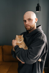 Vertical portrait of handsome bald man man hugging ginger cat looking at camera. Bearded pet owner in black hoodie embracing feline companion, radiating warm domestic connection and tender affection.